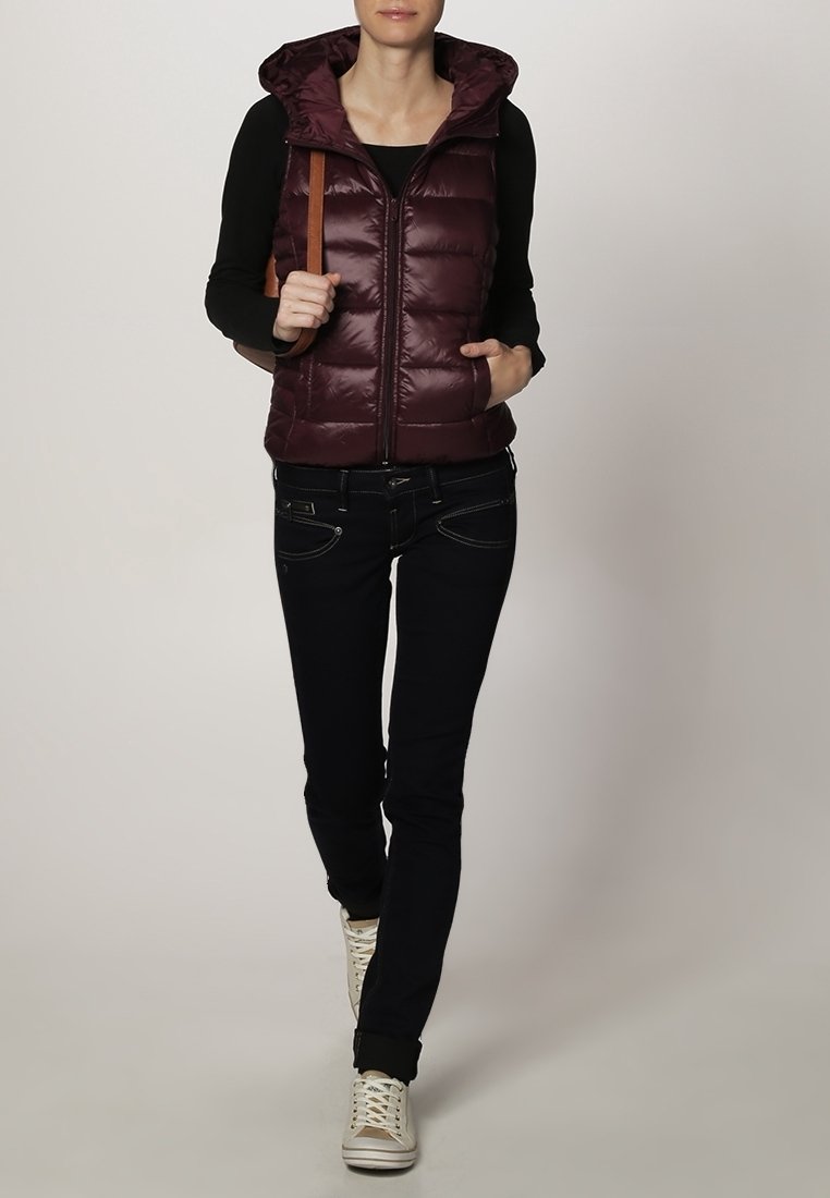 Bordeauxrote Steppweste mit Kapuze und vertikalem Steppmuster, kombiniert mit schwarzer Jeans und weißen Sneakern, mit einem beigen Schulterriemen.