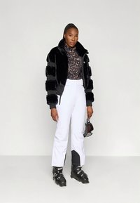 Czarna kurtka z sztucznego futra z poziomymi paskami, zestawiona z brązową bluzką w cętki lamparta i białymi spodniami narciarskimi. Czarne buty narciarskie dopełniają strój.