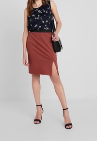Donna che indossa un top senza maniche floreale blu navy, una gonna color ruggine con spacco sulla coscia, tacchi neri con cinturino alla caviglia e porta una borsa a tracolla nera.