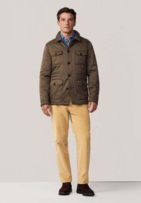 Chaqueta marrón acolchada con cuatro bolsillos frontales, botones negros y cuello, llevada sobre una camisa azul y combinada con pantalones beige y zapatos oscuros.
