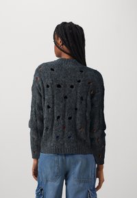 Personne aux cheveux tressés portant un pull en maille gris foncé usé avec des trous, associé à un pantalon cargo en denim bleu, vue de dos.