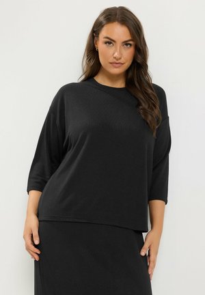 Vrouw met lang bruin haar die een losse zwarte top met driekwart mouwen en bijpassende rok draagt, staande tegen een effen witte achtergrond.