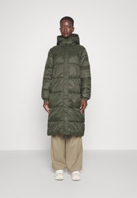 Lang, puffy olivengrøn frakke med hætte, der har en frontlynlås og elastiske manchetter, iført over lyse khaki bukser og hvide sneakers.