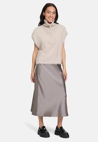 Frau trägt einen beigen, flauschigen, kurzärmligen Pullover, einen grauen Satin-Midirock und schwarze klobige Loafer, steht und lächelt vor einem weißen Hintergrund.