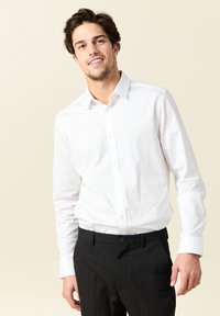 Camisa blanca de manga larga con cuello abotonado. Llevada con pantalones negros ajustados, que cuentan con bolsillos y un tejido texturizado.