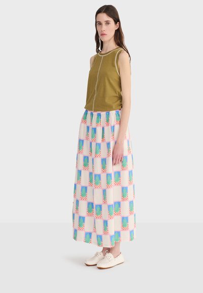 Jeune femme aux longs cheveux bruns portant un haut olive sans manches, une jupe longue pastel à motifs et des baskets blanches, se tenant devant un fond uni.