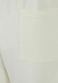 Cremefarbene Hose mit glatter Textur, ausgestattet mit einer quadratischen Gesäßtasche und feinen Nähdetails.