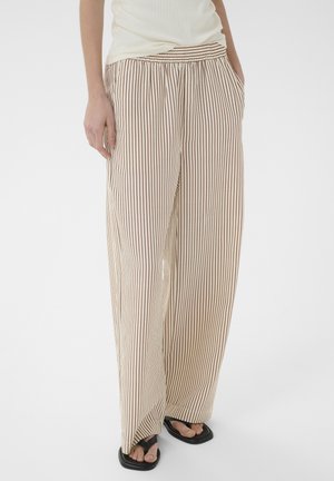 Persoană purtând pantaloni largi bej cu dungi verticale subțiri maro, top alb şi sandale negre cu vârf pătrat, stând cu mâinile în buzunare.