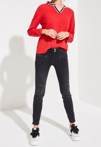 Rød langærmet top med en sort og hvid V-hals, parret med sorte skinny jeans med lynlåse og et slankt design, iført hvide sneakers.