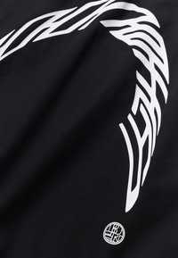 Λευκό αφηρημένο κυρτό γραφικό και μικρό κυκλικό λογότυπο σε μαύρο ύφασμα, που δείχνει πτυχώσεις και υφή υφάσματος.