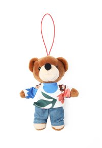Plyšový medvídek jako přívěšek na klíče s hnědou kožešinovou tváří, oblečený v barevném květinovém tričku a modrých džínových kalhotách, má červenou zavěšovací smyčku.