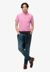 Lyserød polo skjorte, denim jeans og burgundy sko. Ternet skjorte bundet om taljen. Ur på håndleddet, står med hænderne i lommerne.
