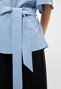 Ανοιχτό μπλε πλεκτό μπλουζάκι με δέσιμο στη μέση, κοντά μανίκια και ευθεία φορά. Φέρει μια διακριτική χρυσή λογότυπο στη κάτω πλευρά.