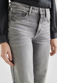 Nahaufnahme einer Person in hochgeschnittenen, ausgewaschenen grauen Jeans und einem schwarzen Langarmshirt, die Hände entspannt an den Seiten.