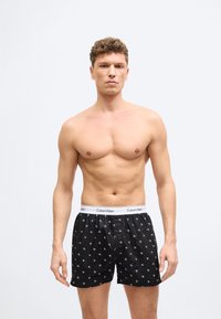 Άνδρας χωρίς μπλούζα με κοντά σγουρά μαλλιά φοράει μαύρα μποξεράκια Calvin Klein με λευκή ζώνη μέσης με λογότυπο, στέκεται μπροστά σε απλό φωτεινό φόντο.