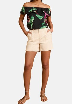 Femme portant un haut noir fleuri épaules dénudées, un short beige taille haute avec poches, et des sandales plates dorées à brides, debout les mains dans les poches.