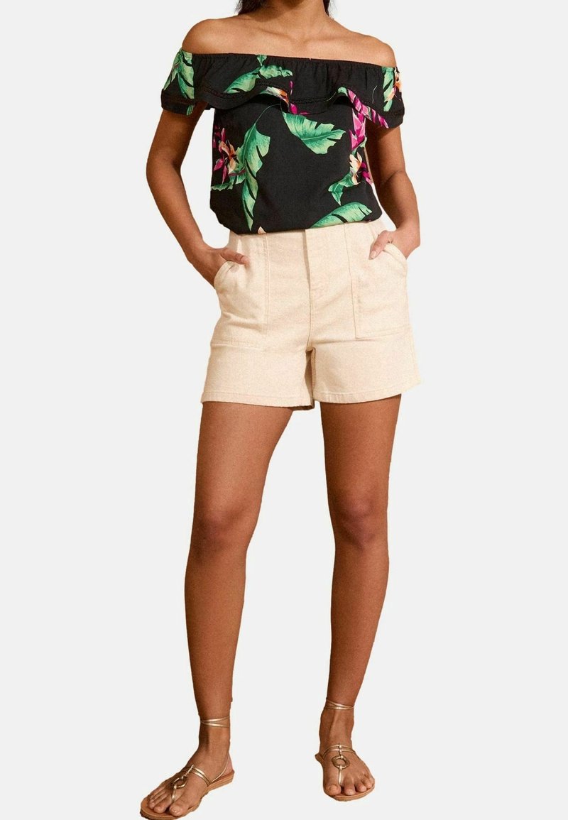 Femme portant un haut noir fleuri épaules dénudées, un short beige taille haute avec poches, et des sandales plates dorées à brides, debout les mains dans les poches.