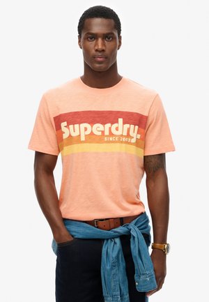 Camiseta melocotón con un gráfico de rayas rojas y naranjas que incluye el texto 'Superdry'. Combinada con jeans oscuros y una camisa de mezclilla azul anudada a la cintura.