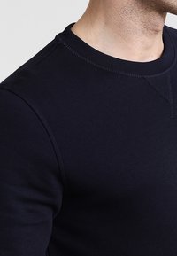 Nahaufnahme der oberen Brust und des Nackens einer Person, die einen dunkelblauen Rundhalsausschnitt-Sweatshirt mit sichtbaren Nähdetails trägt.