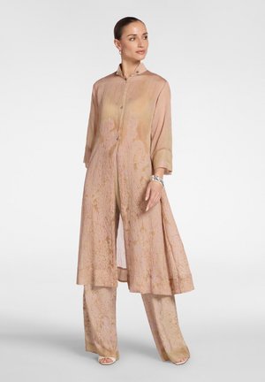 Frau trägt einen fließenden, beige gemusterten langen Mantel und passende weite Hosen, steht vor einem schlichten weißen Hintergrund.
