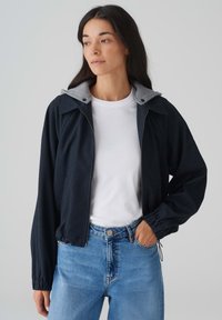Veste navy avec capuche grise, fermeture éclair à l'avant et poignets élastiques. Associée à un t-shirt blanc à col rond et un jean bleu clair. Texture lisse.