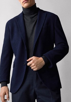 Mand iført mørkeblå blazer over kulgrå rullekravesweater med matchende mørke bukser, justerer blazer med højre hånd.