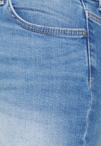 Světle modré džíny z blízkého pohledu, ukazující přední kapsu s žlutým prošíváním a detaily kovových nýtků.