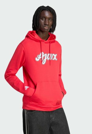 Persoon met dreadlocks die een felrode hoodie draagt met de tekst "Ajax" en een zwarte broek, staand met handen in de voorkant zak.