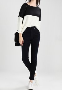 Donna che indossa un maglione a blocchi di colore bianco e nero, jeans skinny neri a vita alta, sneakers bianche e porta una borsa a tracolla nera texturizzata.
