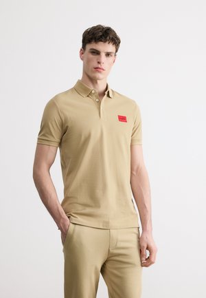 Jeune homme portant un polo beige et un pantalon assorti avec un patch logo rouge sur la poitrine, debout devant un fond blanc uni.