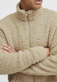 Beige fleecejakke med høy krage, full glidelås og teksturert stoff. Mansjettene og kroppen har et mykt, pelst jakt utseende.