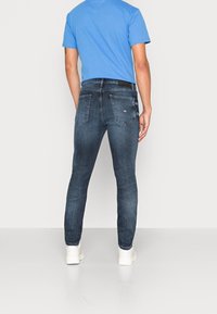 Jeans pitillo azul oscuro con acabado desgastado, dos bolsillos traseros y un parche de cuero con marca. Combinados con una camiseta azul claro y zapatillas blancas.