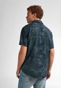 Camisa de manga corta, de color verde oscuro, con un estampado de hojas de palma, que presenta un cuello y un dobladillo recto, combinada con unos vaqueros de mezclilla de color azul claro.