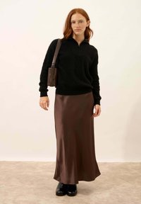 Zwart gebreid pullover met een ritssluiting in de hals, gecombineerd met een bruine satijnen maxi rok en zwarte platformlaarzen. Een kleine bruine schoudertas wordt gedragen.