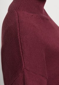 Pull en maille bordeaux avec un col haut à côtes, un design de points texturés et des coutures d'épaule abaissées ; tissu doux et léger.