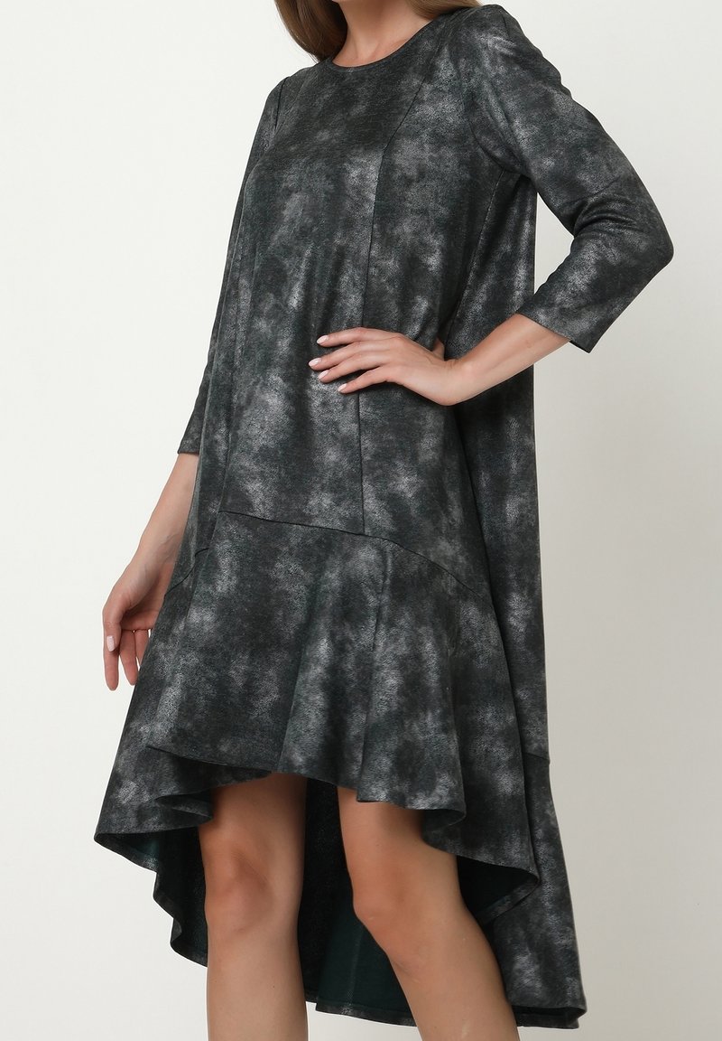 Femme portant une robe à manches longues avec un ourlet asymétrique en tissu gris foncé moucheté, main posée sur la hanche, sur un fond clair uni.