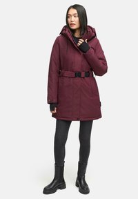Bordeaux geïsoleerde parka met een riem, een verstelbare capuchon en duimopeningen, gecombineerd met zwarte skinny jeans en dikke zwarte laarzen.