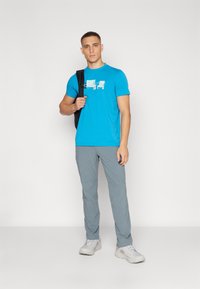 Camiseta azul con diseño gráfico, pantalones grises y zapatillas grises. El modelo lleva una mochila negra y está de pie sobre un fondo sencillo.