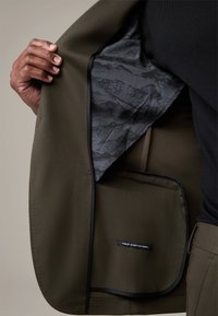 Olivgrüne Jacke mit strukturiertem Oberfläche; Innenfutter hat ein schwarz-graues Muster. Enthält ein verstecktes Etikett für die Stoffqualität.