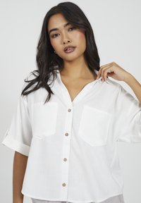 Młoda kobieta z długimi ciemnymi włosami nosi luźną białą koszulę zapinaną na guziki, trzymając kołnierz jedną ręką na jednolitym jasnym tle.