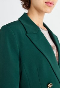 Gros plan sur une personne portant un blazer vert foncé avec des revers larges et un bouton doré, par-dessus une chemise à motifs.