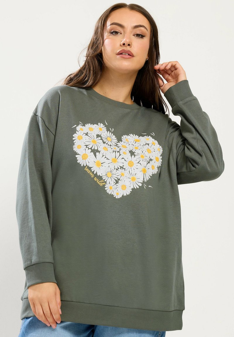 Junge Frau richtet ihre Haare, trägt ein übergroßes grünes Sweatshirt mit weißem Gänseblümchen-Herz-Design und dem Schriftzug "bloom wild", kombiniert mit blauen Jeans.