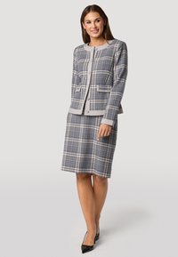 Marineblauw en grijs geruite kostuumset met een knoopblazer, twee voorzakken en een rok tot op de knie. Kenmerkt zich door een ronde halslijn en een soepele textuur.