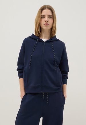 Jeune femme aux cheveux blonds et raides portant un sweat à capuche zippé bleu marine et un pantalon assorti, les mains dans les poches.