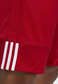 Shorts de sport rouges en tissu texturé, présentant trois rayures blanches sur le côté près de l'ourlet, avec une coupe décontractée.