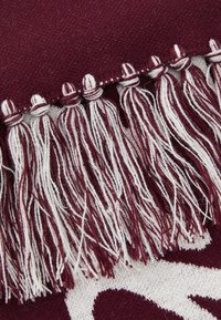 Tecido bordeaux com detalhes de franjas brancas. O tecido tem uma textura suave, apresentando um padrão tecido e franjas decorativas ao longo da borda.