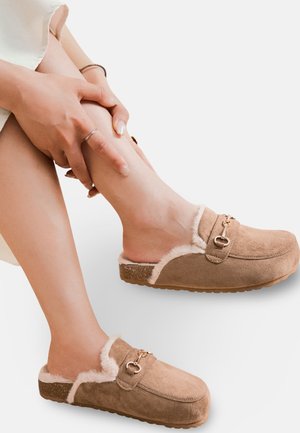 Sabots en suède marron avec doublure en fausse fourrure, design à enfiler et accents en métal doré. Semelle en caoutchouc texturé pour une meilleure adhérence.