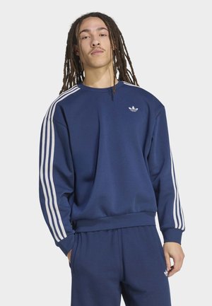 Giovane uomo con lunghi dreadlocks che indossa una tuta Adidas blu scuro con strisce bianche e loghi, in piedi davanti a uno sfondo chiaro.