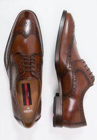 Scarpe brogue in pelle marrone con perforazioni dettagliate, un classico design con lacci, texture liscia e accenti neri a contrasto sul tallone.