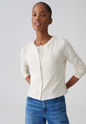 Donna dalla pelle scura che indossa un cardigan bianco con bottoni e jeans blu, in piedi con le mani dietro la schiena su uno sfondo semplice.